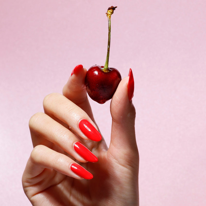 A hand with long, bright red LONDONTOWN Maraschino Cherry nails holds a single cherry by its stem against a light pink background.
