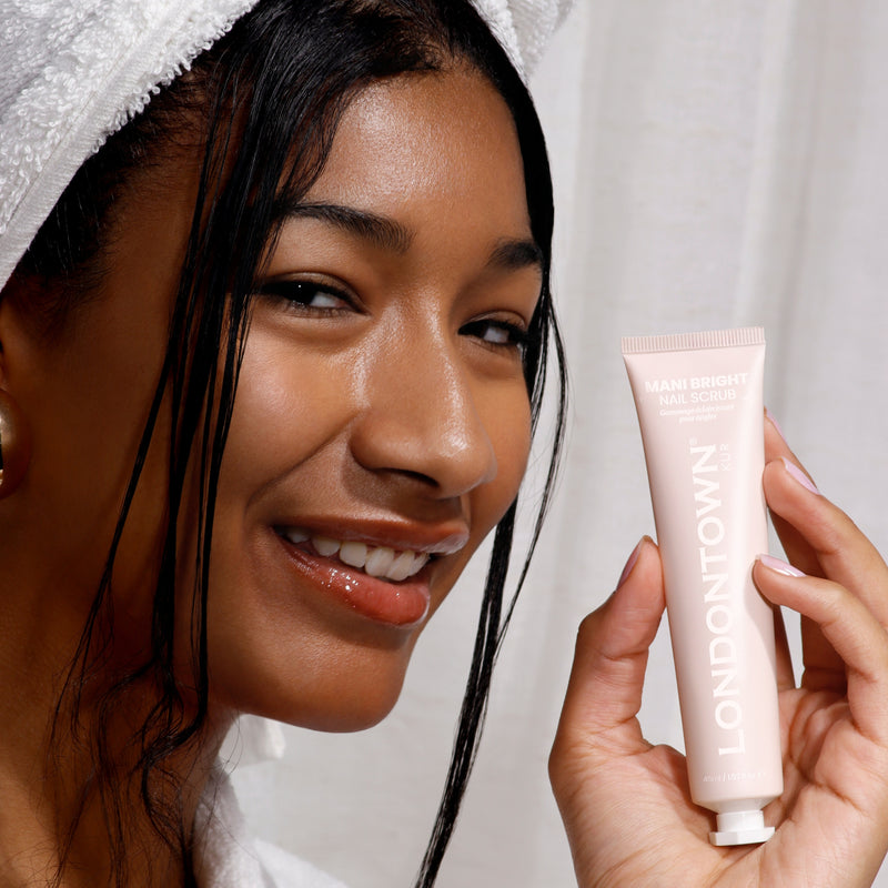 A person in a towel proudly displays a tube of LONDONTOWN Mani Bright Nail Scrub, smiling confidently because this nail solution effortlessly tackles stained nails.