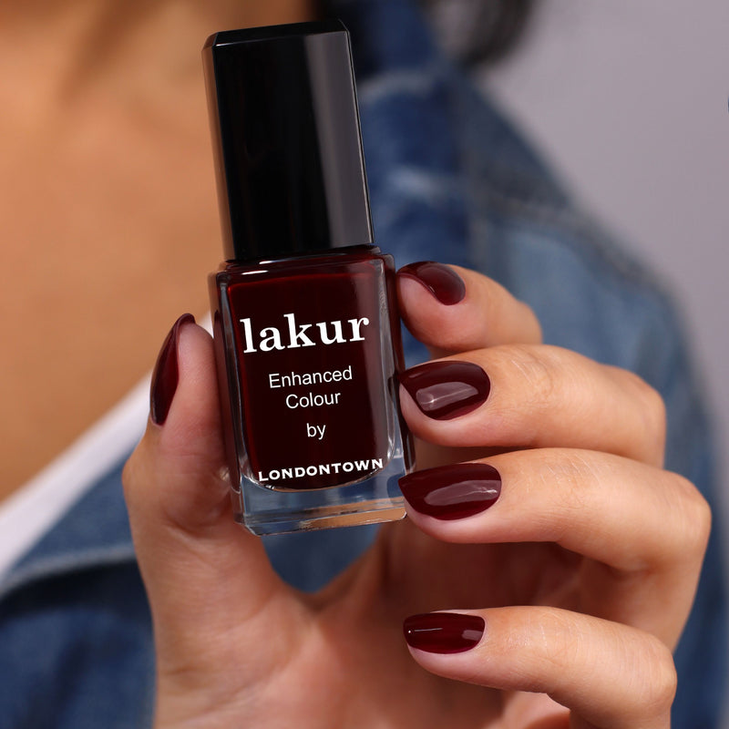 A hand holds a bottle of LONDONTOWN's "Elderberry" nail polish, its captivating blackened purple hue perfectly matching the painted fingernails.