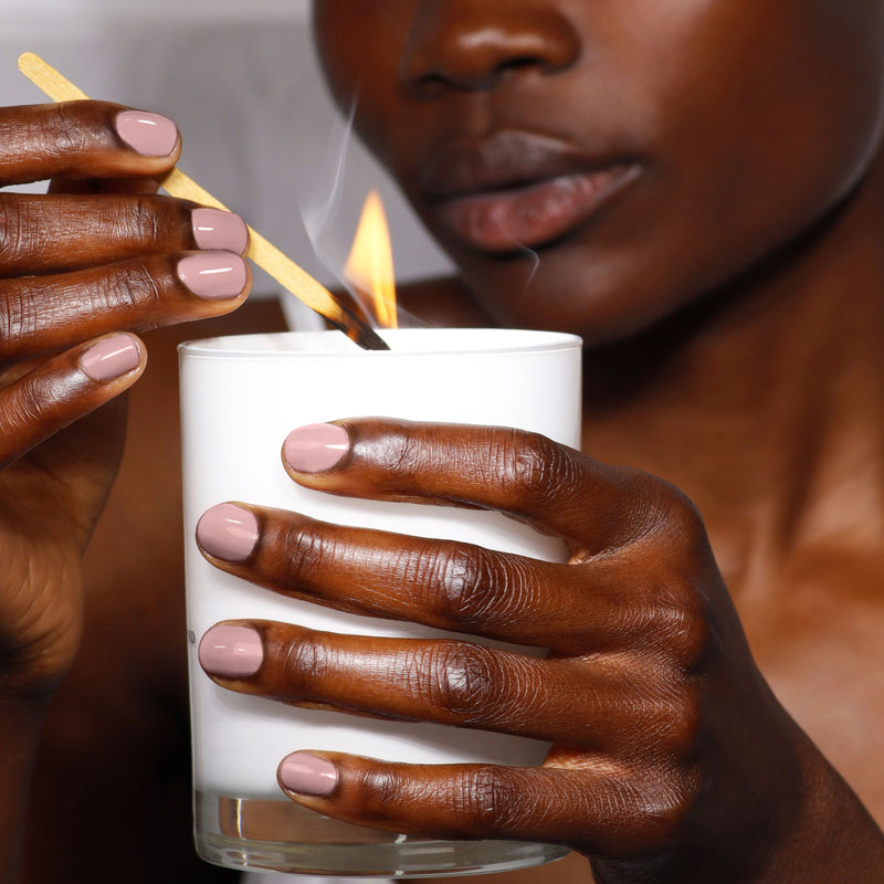 A person with well-groomed nails painted in LONDONTOWN's soft mauve "Do Not Disturb" lights a white candle using a wooden match.