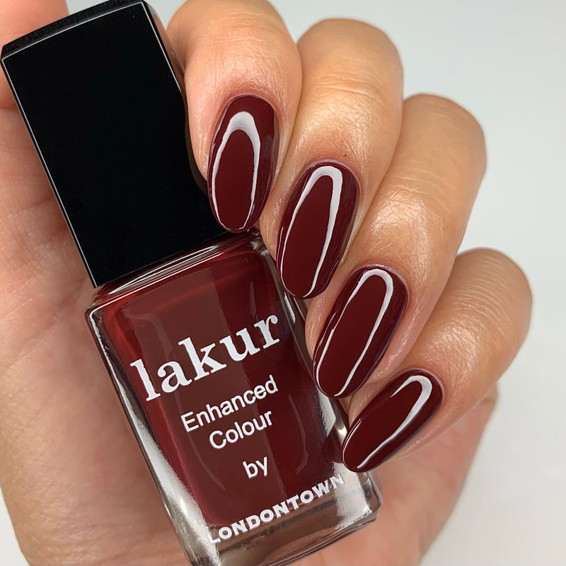 A manicured hand with mysterious blackened purple nails holds a bottle of LONDONTOWN's Elderberry nail polish, showcasing an enchanting depth of dark red color.