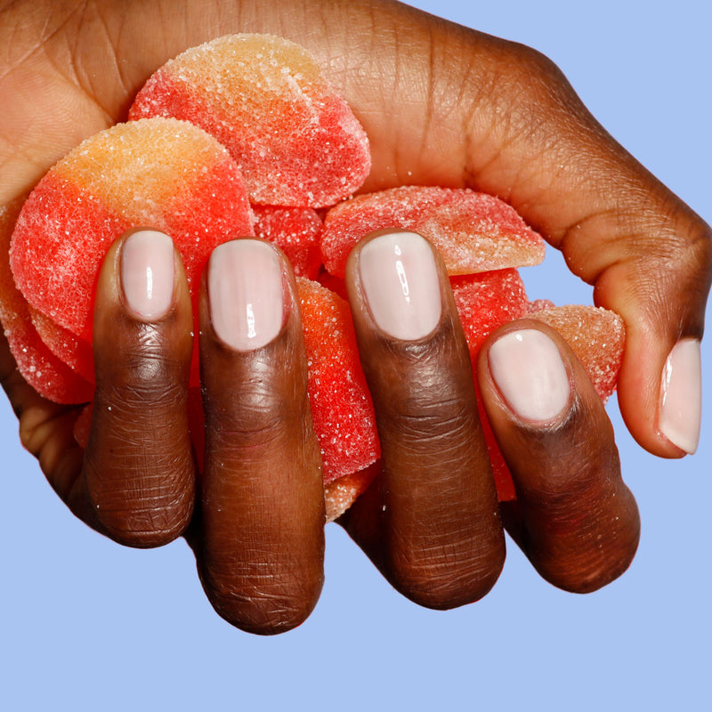 A hand with creamy, manicured nails painted in LONDONTOWN’s Double Scoop shade holds red and orange gummy candies against a light blue background.