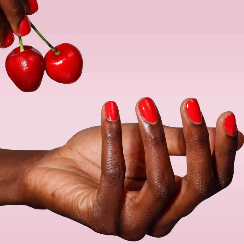 A hand with Maraschino Cherry by LONDONTOWN bright red nails is held open as another hand holds two cherries above it, all set against a pink background.