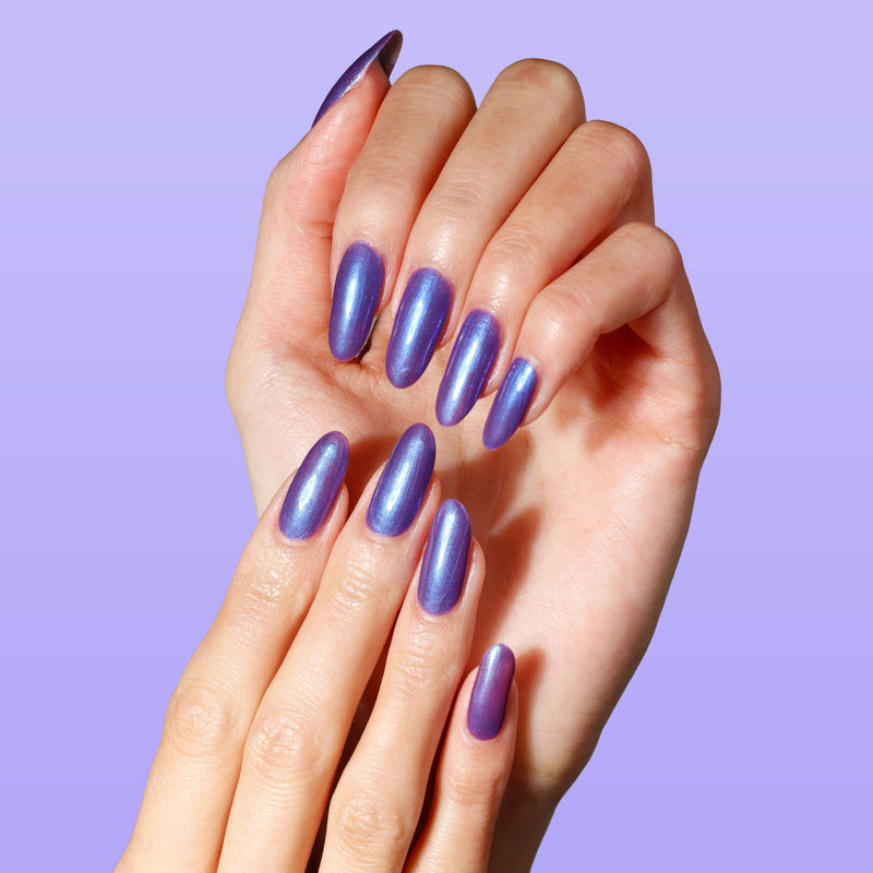Hands with long, almond-shaped nails painted in LONDONTOWN's Wildberry Freeze—a shiny metallic purple polish with a blue shimmer—set against a light purple background.