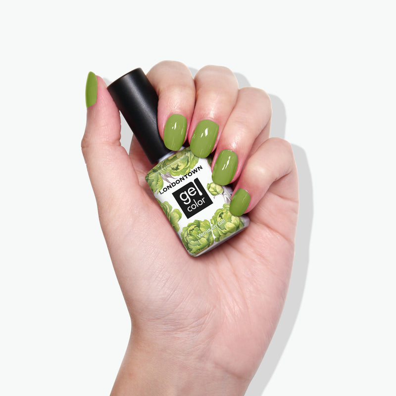 A hand with green painted nails holding a bottle of LONDONTOWN's gel color nail polish in the shade Tipsy Mermaid, featuring a matching green label reminiscent of a vibrant parakeet.