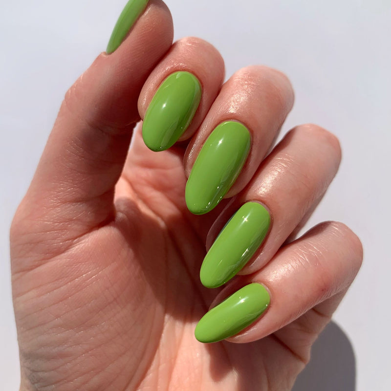 A hand with long, almond-shaped nails painted in LONDONTOWN's glossy, parakeet green "Tipsy Mermaid" nail polish against a plain background.