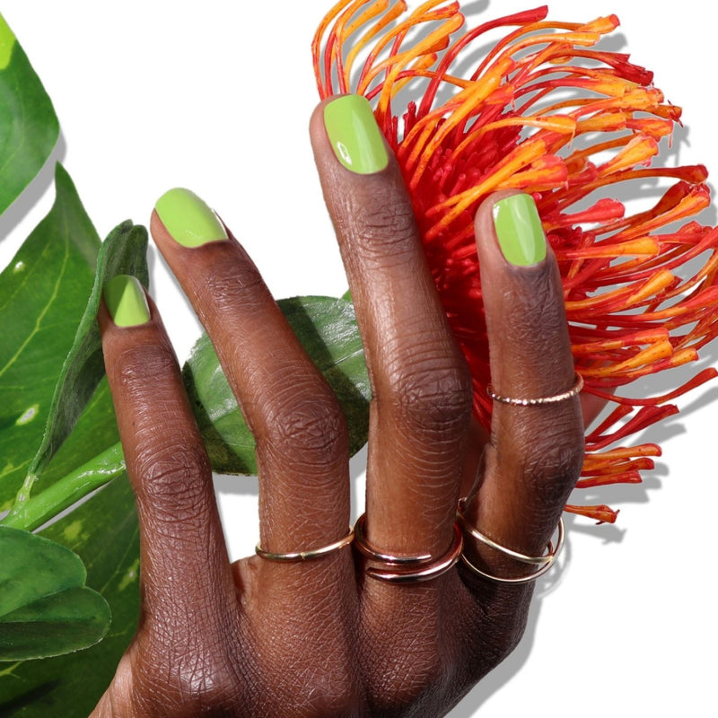 A hand with LONDONTOWN's Tipsy Mermaid green nail polish wears gold rings and holds an orange flower against a white background adorned with green leaves, reminiscent of a parakeet's vibrant plumage.