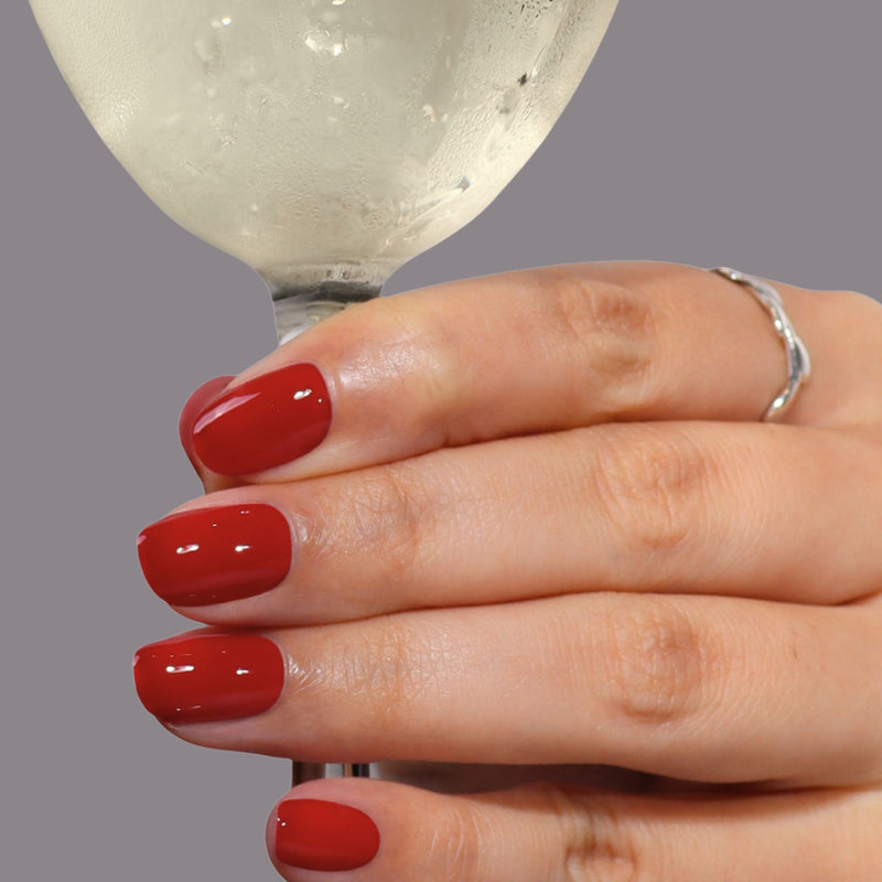 A hand with LONDONTOWN’s Truly Ruby polished nails and a silver ring holds a glass of white wine against a plain gray background.