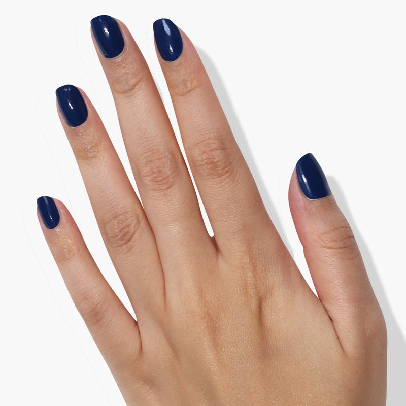 A hand with neatly manicured nails is painted in LONDONTOWN's Under The Stars, a glossy dark blue nail polish reminiscent of vintage denim, against a white background.
