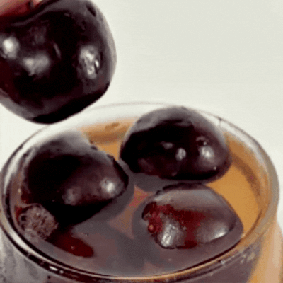 A hand drops a dark plum into a glass of iced tea and plums; the splash evokes the fizzy refreshment of LONDONTOWN’s Cherry Cola Essentials.