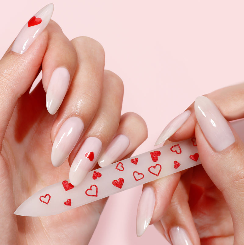 Close-up of hands effortlessly using the LONDONTOWN Heart Glass Nail File, emphasizing damage prevention. Nails feature a light pink manicure with delicate red heart accents.