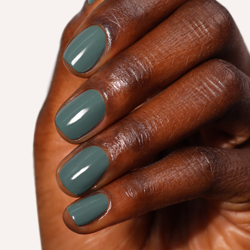 A hand with medium-dark skin tone displays nails painted in a glossy, sumptuous olive green color, Velvet Drapes by LONDONTOWN, against a plain background.