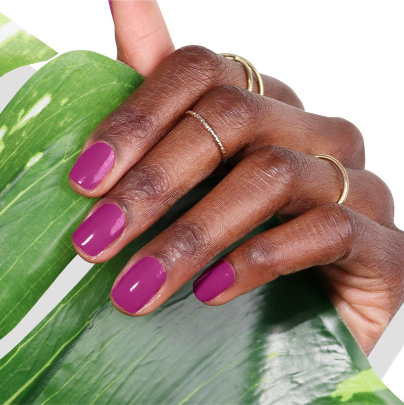 A hand adorned with two rings and painted with LONDONTOWN's Violet Hibiscus nail polish holds a green leaf.