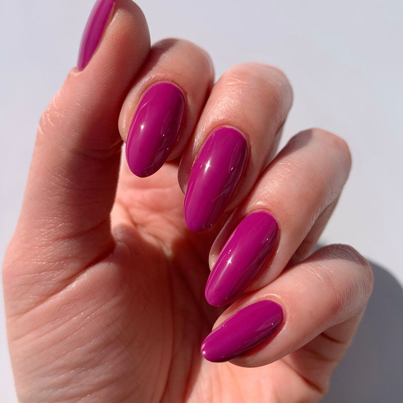 A hand with long, almond-shaped nails painted in LONDONTOWN's glossy Violet Hibiscus polish shines with a deep, ultraviolet allure.