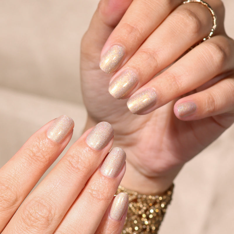 Close-up of hands with short nails painted in LONDONTOWN's "You're Golden" holographic gold polish, accessorized with a gold ring and beaded bracelet.