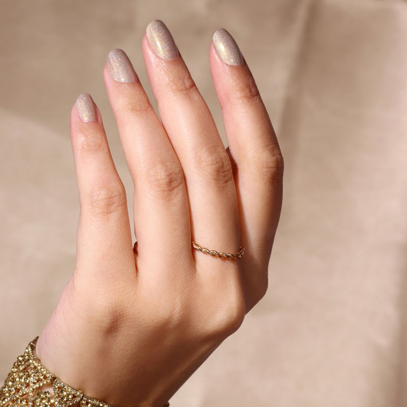 A hand with neatly manicured, You're Golden holographic gold glitter nails by LONDONTOWN is shown wearing a thin, twisted gold ring against a neutral background.