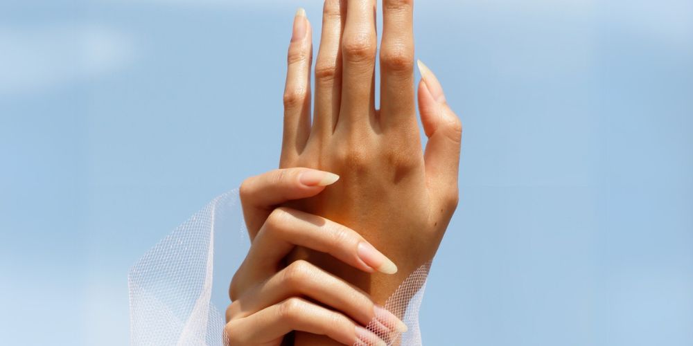 A pair of hands with long nails are held gracefully against a clear blue sky. A sheer white fabric partially covers the wrist.