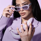 A woman in a lavender blazer and sunglasses holds glossy, lavender bananas against a pastel purple backdrop, capturing the modern style essence of LONDONTOWN's Lavender Illuminating Nail Concealer.