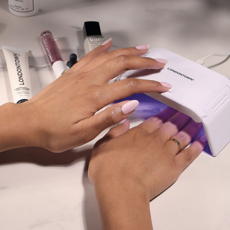 A person cures freshly painted nails with the LONDONTOWN Foldable UV/LED Gel Lamp for a fast manicure, surrounded by nail care products and polish bottles on the table.