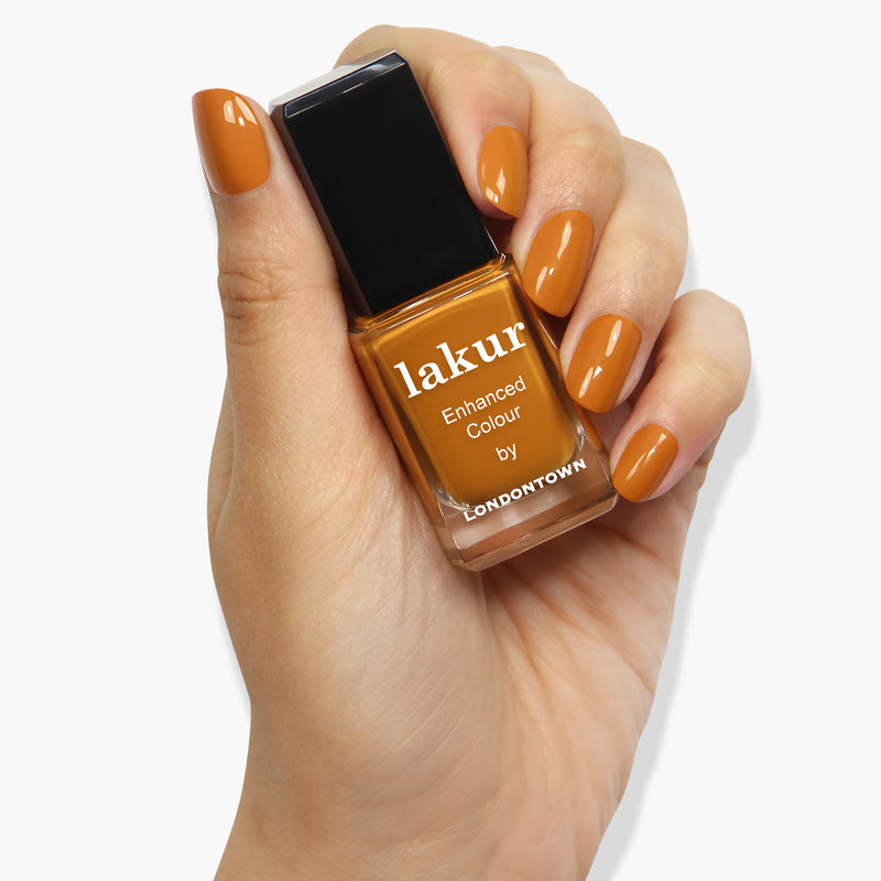 A hand with neatly manicured nails, painted in a matching orange-brown shade akin to robust goldenrod, holds a bottle of "Bee's Knees by LONDONTOWN" nail polish in the same color.