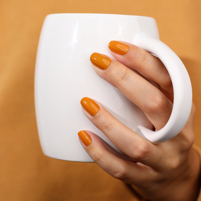 A hand adorned with LONDONTOWN's Bee's Knees orange nail polish holds a white coffee mug against a robust goldenrod background.