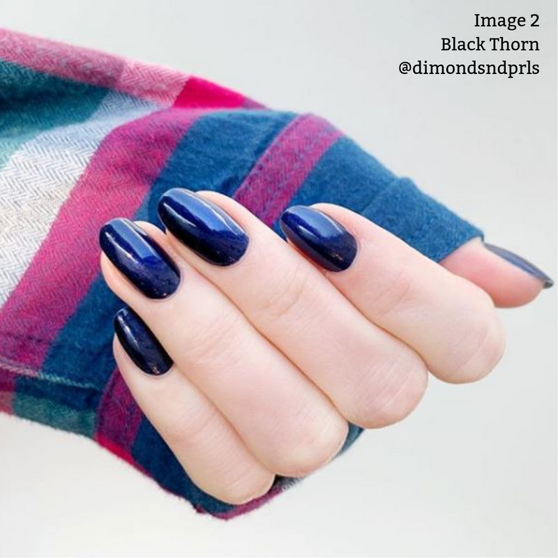 A hand with long, oval-shaped nails painted in a dark, glossy shade of Black Thorn by LONDONTOWN. The person is wearing a multi-colored, striped long-sleeve shirt. Text reads: "Image 2 Black Thorn @dimondsndprls" highlighting their vegan cruelty-free formula.