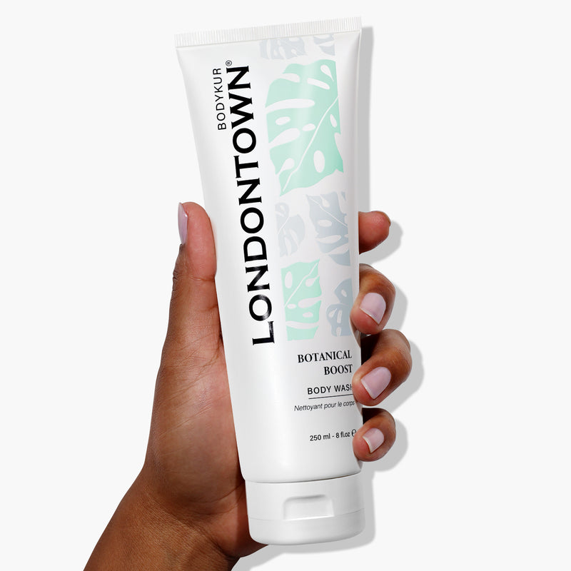 A hand is holding a tube of LONDONTOWN Botanical Boost Body Wash against a white background. The white tube, adorned with black text and green leaf designs, promises to elevate your everyday shower into a moment of luxury.