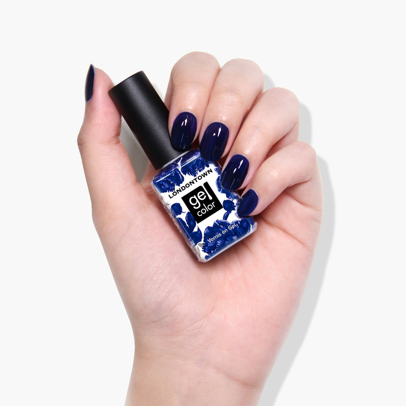 A hand with neatly manicured nails painted in a Buckingham Blue polish is holding a bottle of LONDONTOWN gel color nail polish, perfect for use with a UV LED light. The vegan, cruelty-free formula ensures guilt-free glam.