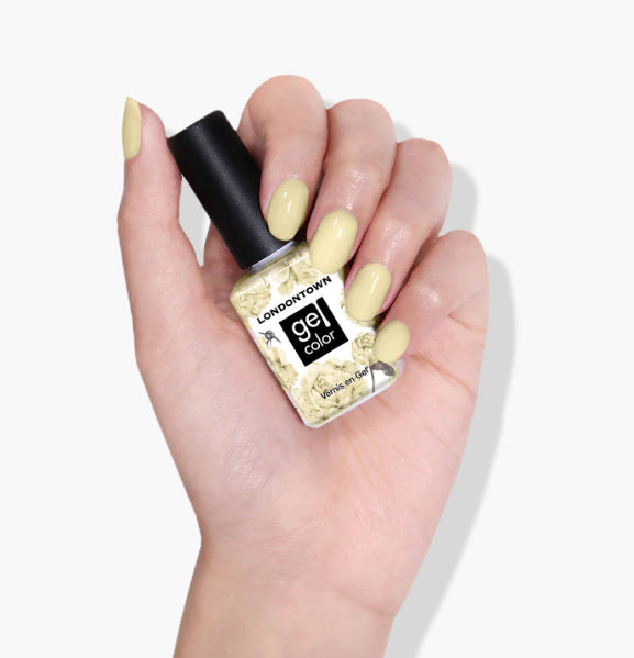 A hand with light yellow painted nails holds a bottle of LONDONTOWN Buttercup UV/LED Gel Color nail polish against a white background, showcasing the pastel yellow creme shade.