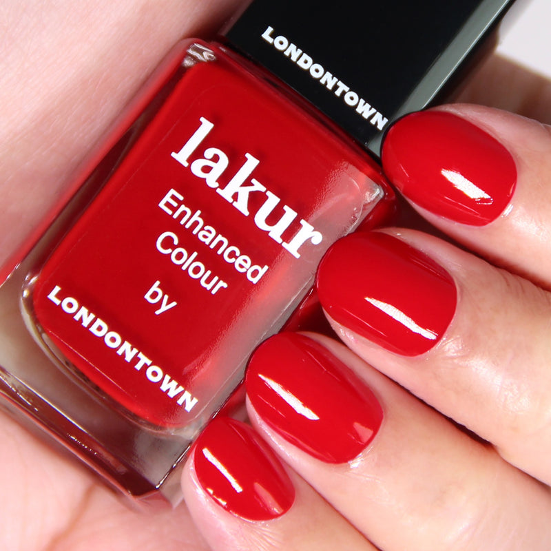 Close-up of a hand with neatly manicured nails painted in a deep red crème, holding a bottle of LONDONTOWN's Changing of the Guards Lakur Enhanced Colour, a vegan and cruelty-free red nail polish.
