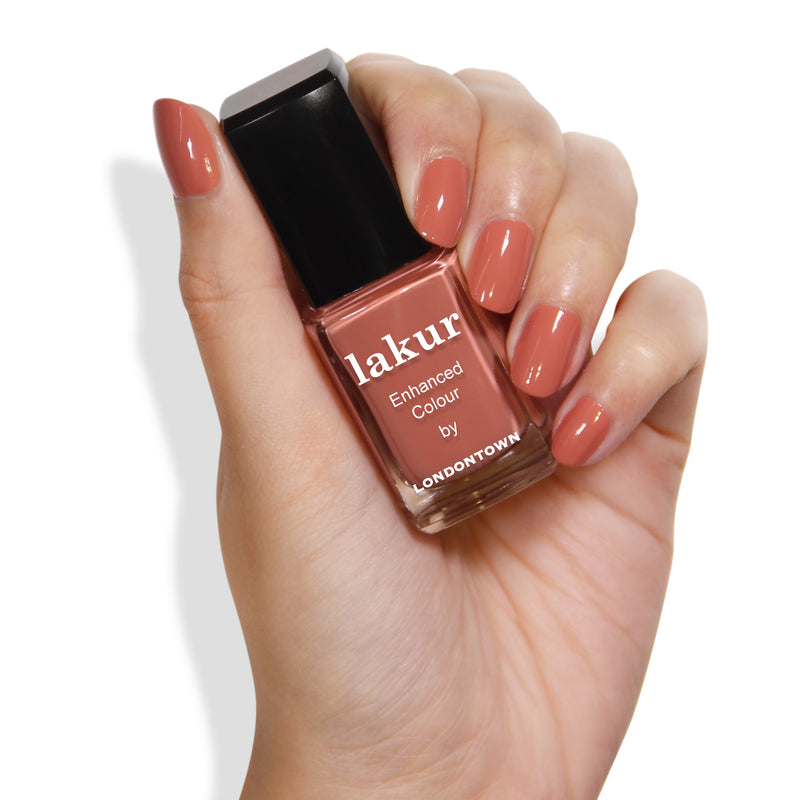 A hand holds a bottle of Cactus Coral vegan nail polish in a lush dusty coral shade with the text "Enhanced Colour by LONDONTOWN" on the label. The nails are painted in a matching color.