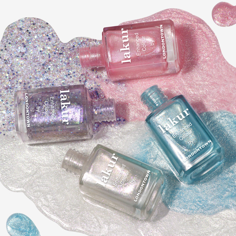 Four bottles of LONDONTOWN's Coconut Crush nail polish, each in different colors, are lying on a mix of pink, blue, and silver shimmering paint drips dusted with ultrafine glitter. The bottle colors are light pink, pink, silver, and light blue.
