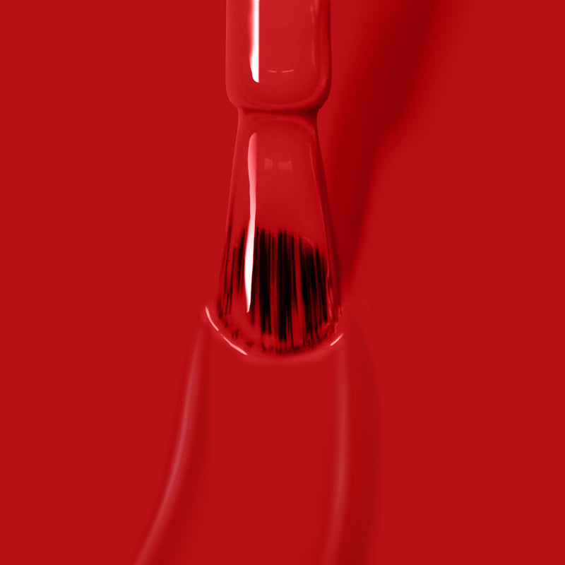 A brush is coated with Changing of the Guards deep red crème paint by LONDONTOWN, set against a red background with visible bristles at the tip blending into the painted area.