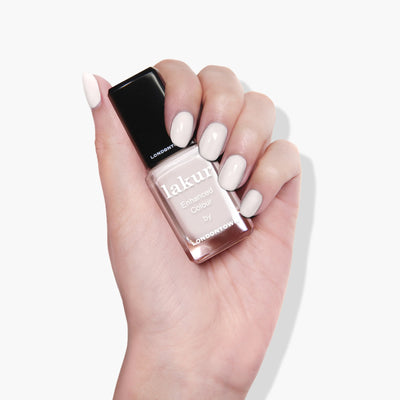 A hand holding a bottle of LONDONTOWN Chelsea Porcelain (Professional) nail polish in a milky off-white crème shade against a white background. This vegan, cruelty-free formula ensures chic nails with an ethical touch.