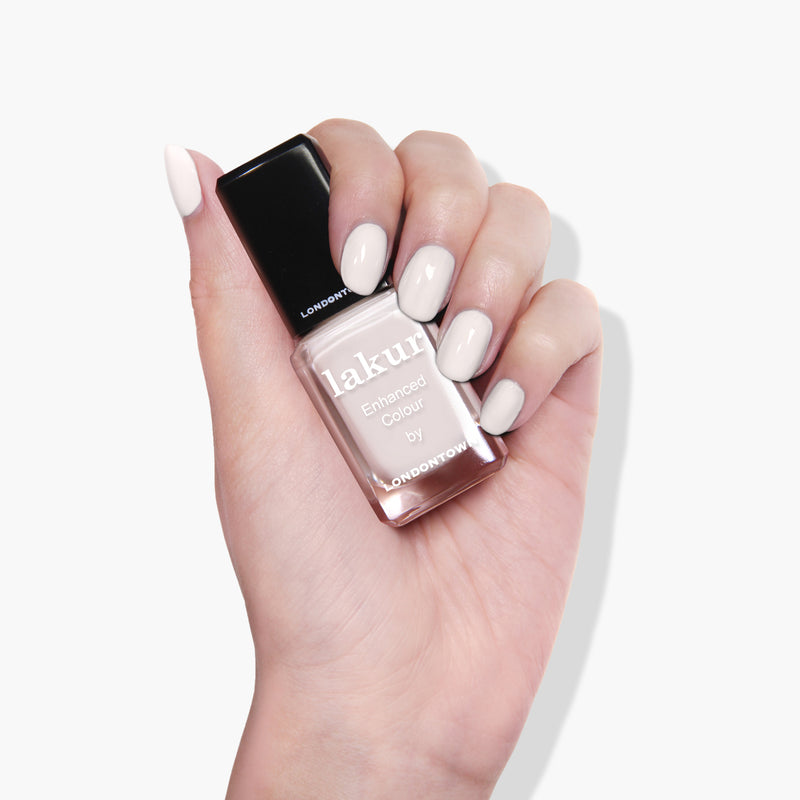 A hand holding a bottle of LONDONTOWN's Chelsea Porcelain cruelty-free nail polish with white polished nails matching the milky off-white crème vegan nail polish color.