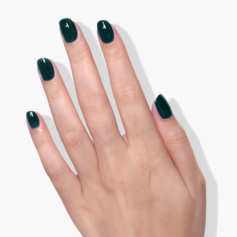 A hand with short, neatly manicured nails painted in LONDONTOWN's Chivvy Along, a dark green vegan nail polish, is shown against a plain white background.