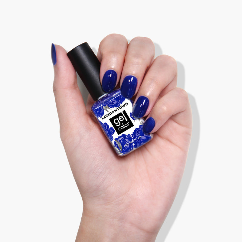 A hand with blue-painted nails holds a bottle of LONDONTOWN Gel Color nail polish in the stunning "Cocktail Hour" purple crème shade.
