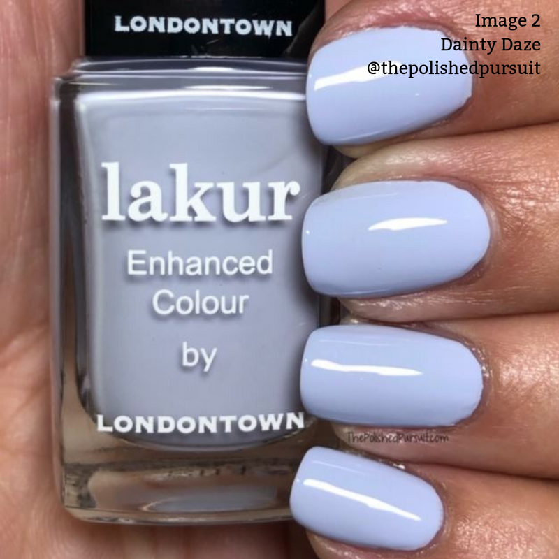 A hand with neatly manicured nails painted in a pale lavender shade holds a bottle of LONDONTOWN's vegan, cruelty-free Lakur Enhanced Colour nail polish in the color Dainty Daze.