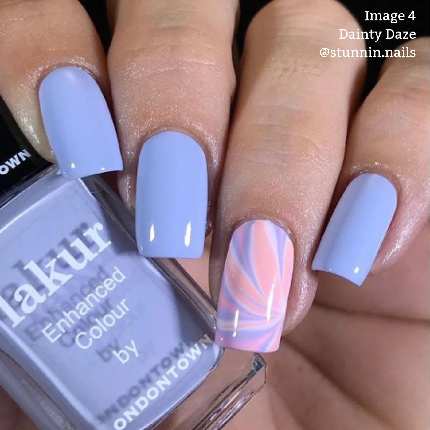 Close-up of a hand with four fingernails painted in a light lavender shade. The ring fingernail features a pink and white swirl design. A bottle of LONDONTOWN's Dainty Daze (Professional) vegan, cruelty-free nail polish in matching lavender is held between the thumb and index finger.