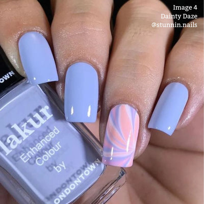 Close-up of a hand with four fingernails painted in a light lavender shade. The ring fingernail features a pink and white swirl design. A bottle of LONDONTOWN's Dainty Daze (Professional) vegan, cruelty-free nail polish in matching lavender is held between the thumb and index finger.