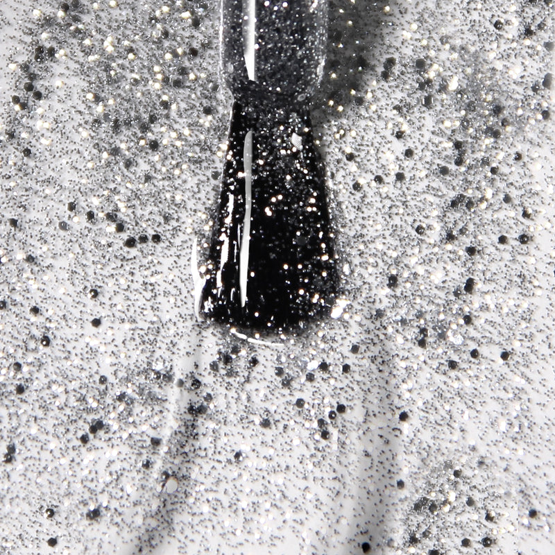Close-up of a black nail polish brush with glitter, creating a speckled pattern on a white surface. The high-shine formula of LONDONTOWN's Diamond Jubilee offers vibrant color and is vegan and cruelty-free.