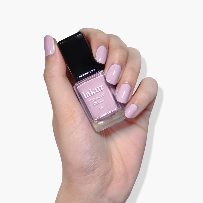 A hand with manicured nails holds a bottle of LONDONTOWN Dolly Mix, a light purple lilac nail polish that's vegan, cruelty-free, and 16+ free.