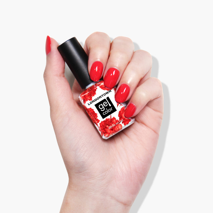 Hand holding a white bottle of LONDONTOWN Down to Dilly gel color polish with a black cap, decorated with red flowers. The nails are painted in a matching glossy red color, cured under UV/LED light.