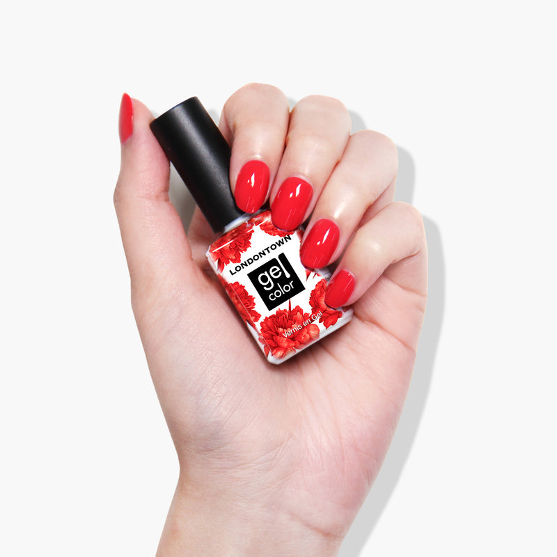 Hand holding a white bottle of LONDONTOWN Down to Dilly gel color polish with a black cap, decorated with red flowers. The nails are painted in a matching glossy red color, cured under UV/LED light.