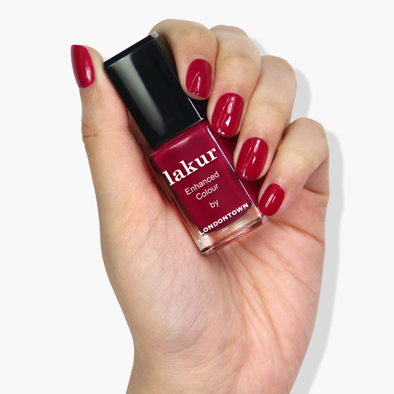 A hand with neatly manicured nails painted in deep red holds a bottle of LONDONTOWN's Dragon Fruit lakur enhanced colour nail polish, known for its cruelty-free and vegan formula.