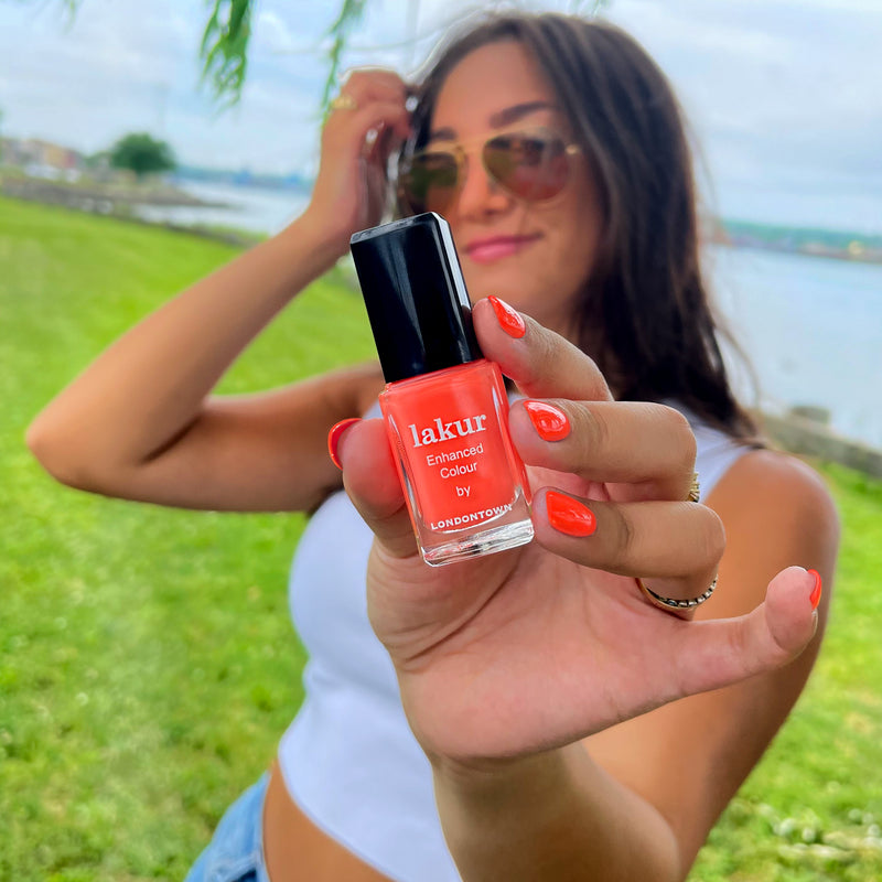 A person outdoors holds up a bottle of LONDONTOWN Flashback nail polish in radiant sunset orange while proudly displaying their vegan cruelty-free painted nails.