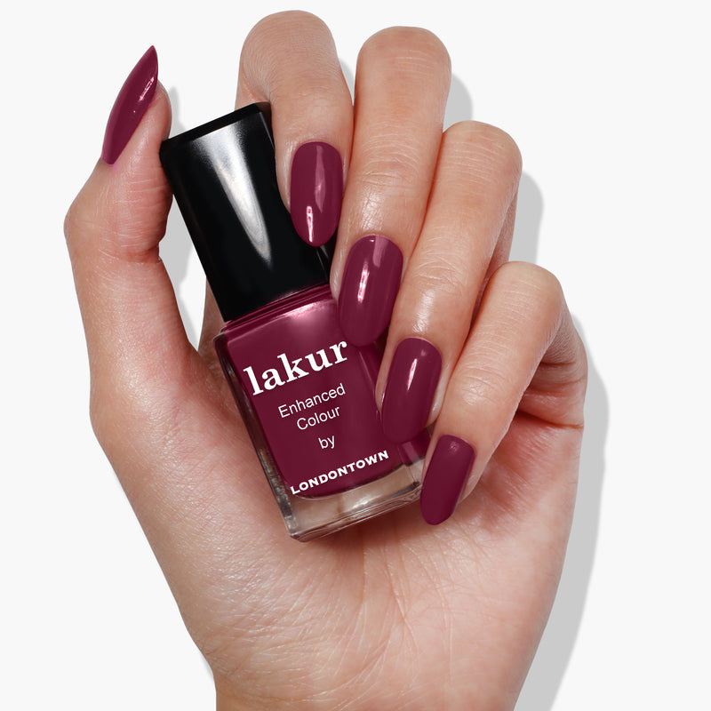 A hand with manicured nails holds a bottle of LONDONTOWN Flirty & Thriving nail polish in a rich, ripened raspberry hue.