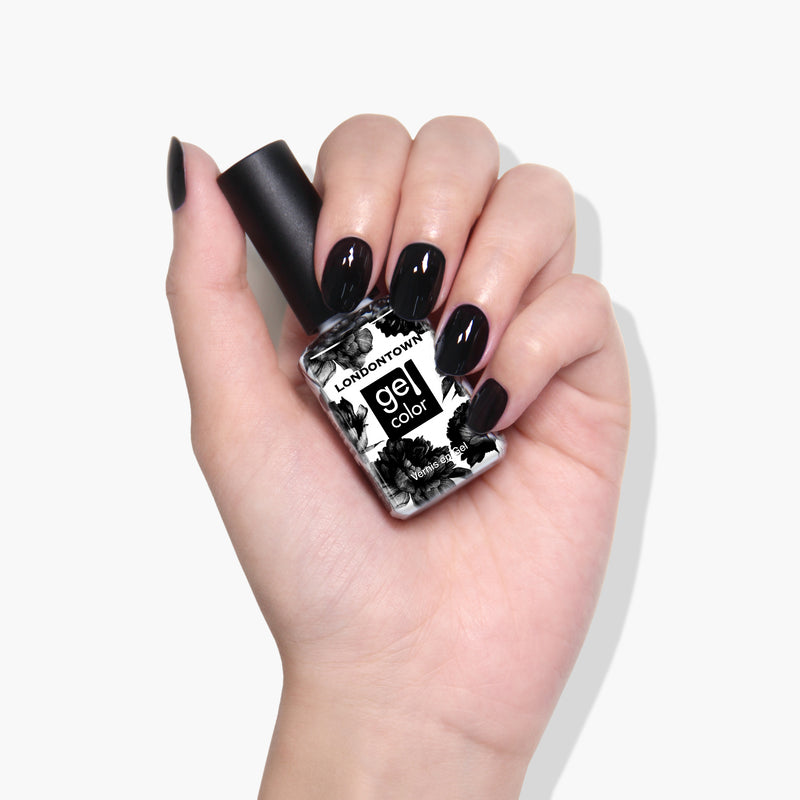 A hand with manicured Chim Cher-ee jet black crème nails holds a bottle of LONDONTOWN Gel Color nail polish against a plain white background.