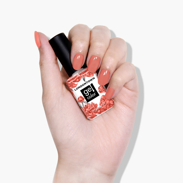 A hand with manicured nails holds a bottle of LONDONTOWN Cactus Coral Gel Color nail polish in a lush, dusty coral shade adorned with floral designs.