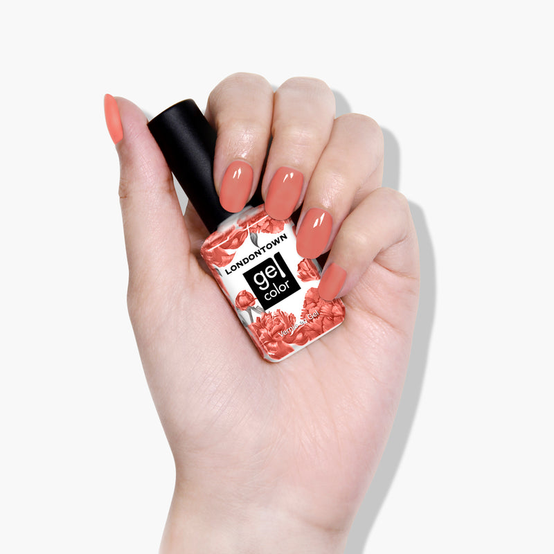 A hand with manicured nails holds a bottle of LONDONTOWN Cactus Coral Gel Color nail polish in a lush, dusty coral shade adorned with floral designs.